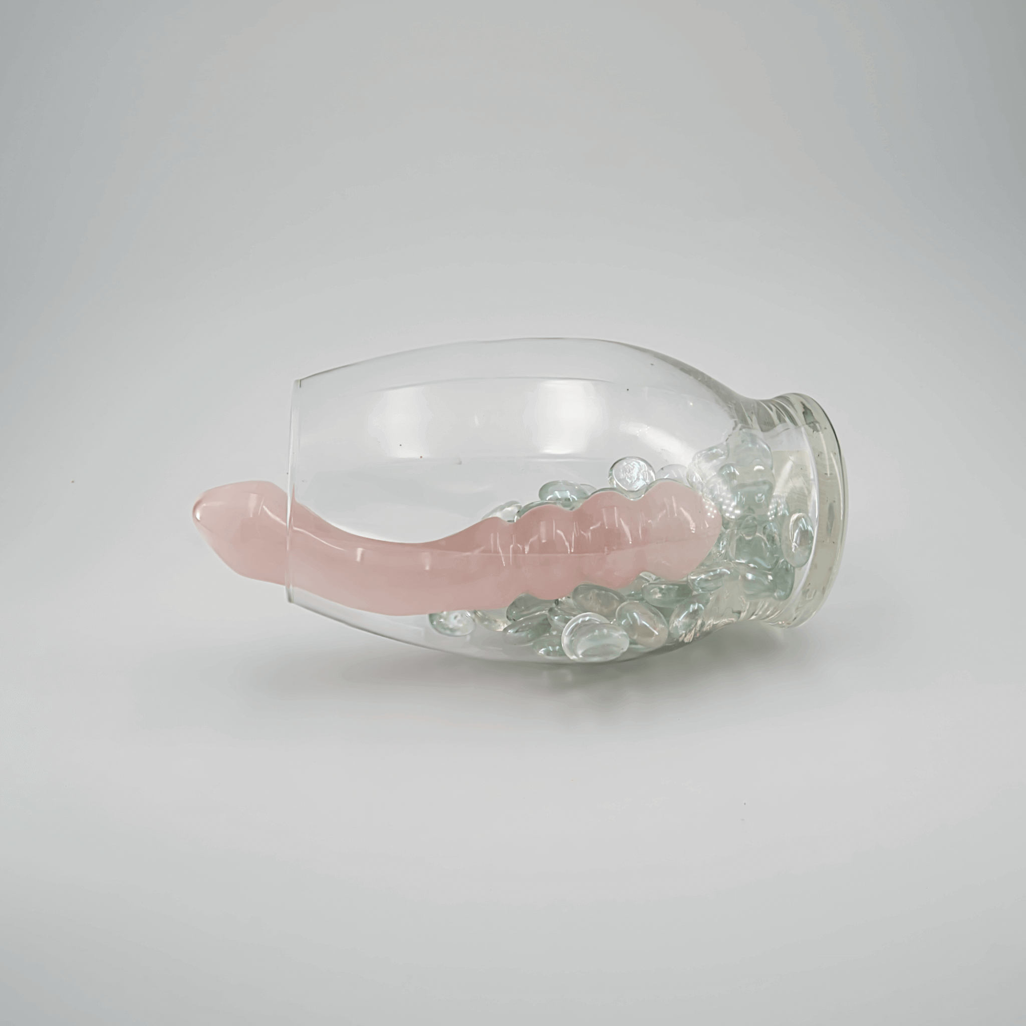 A pink crystal dildo lying inside a glass jar filled with decorative stones, showcasing its elegant design.
